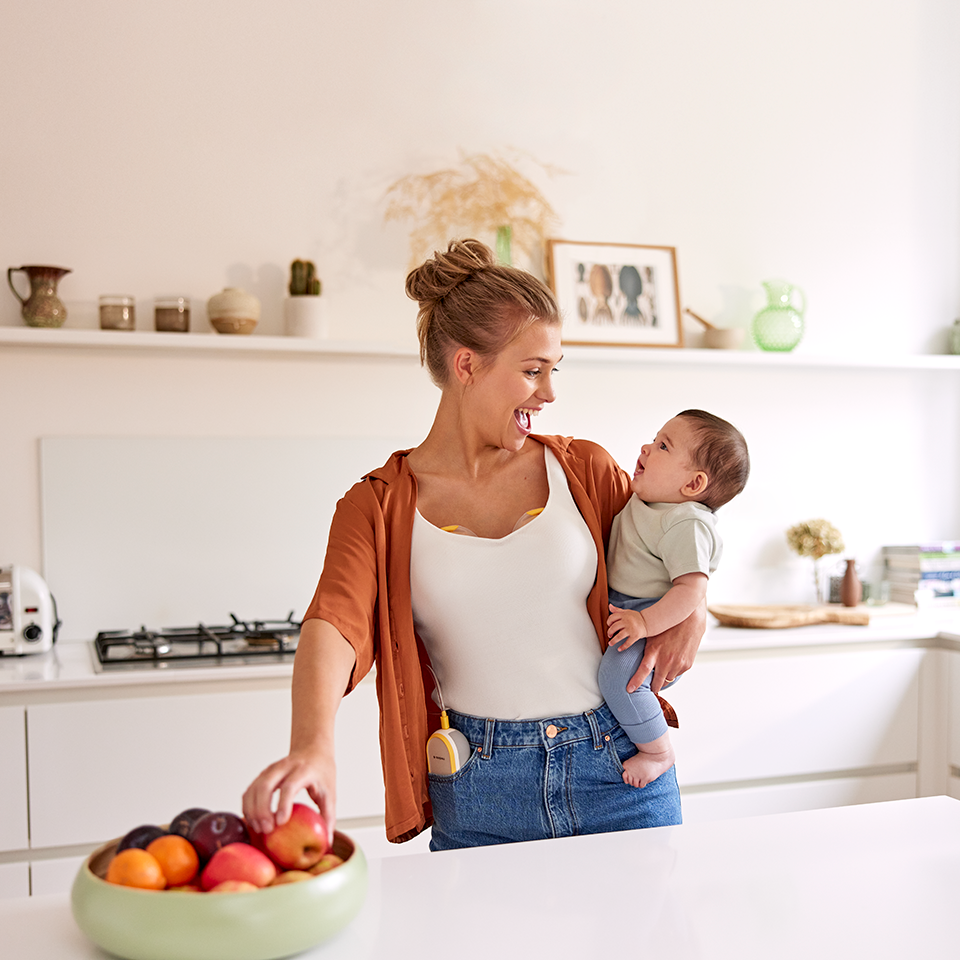 A mom holds her baby and has a snack while wearing her Medela Freestyle™ Hands-free double electric breast pump.