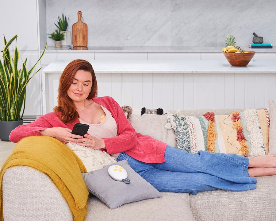 Mam gebruikt haar telefoon terwijl ze uitrust op een bank en haar Medela Solo™ Hands-free enkele elektrische borstkolf gebruikt