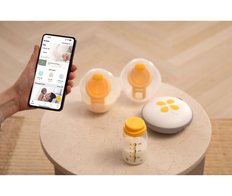 Medela Swing Maxi™ double electric breast pump on table with two Hands-free collection cups and a Medela storage container while mom looks at the Medela app on her phone..