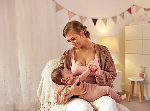 Mamma che indossa un tiralatte elettrico doppio Medela Swing Maxi™ mentre tiene in braccio il suo bambino nella stanza dei bambini