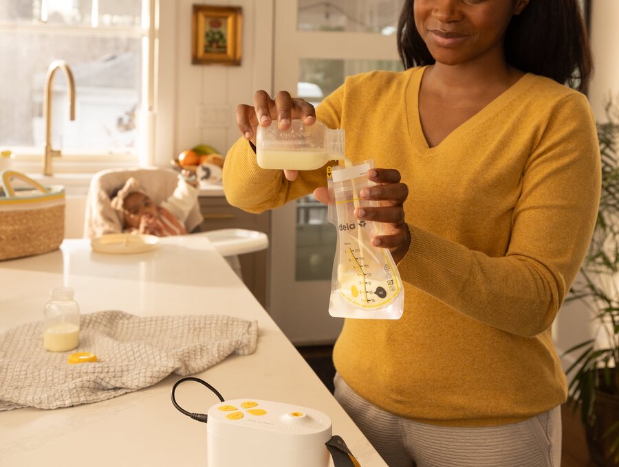 Woman in kitchen pouring breast milk expressed with Pump In Style Pro breast pump into Easy Pour Breast Milk Storage Bag.  Baby is sitting in high chair watching mom.