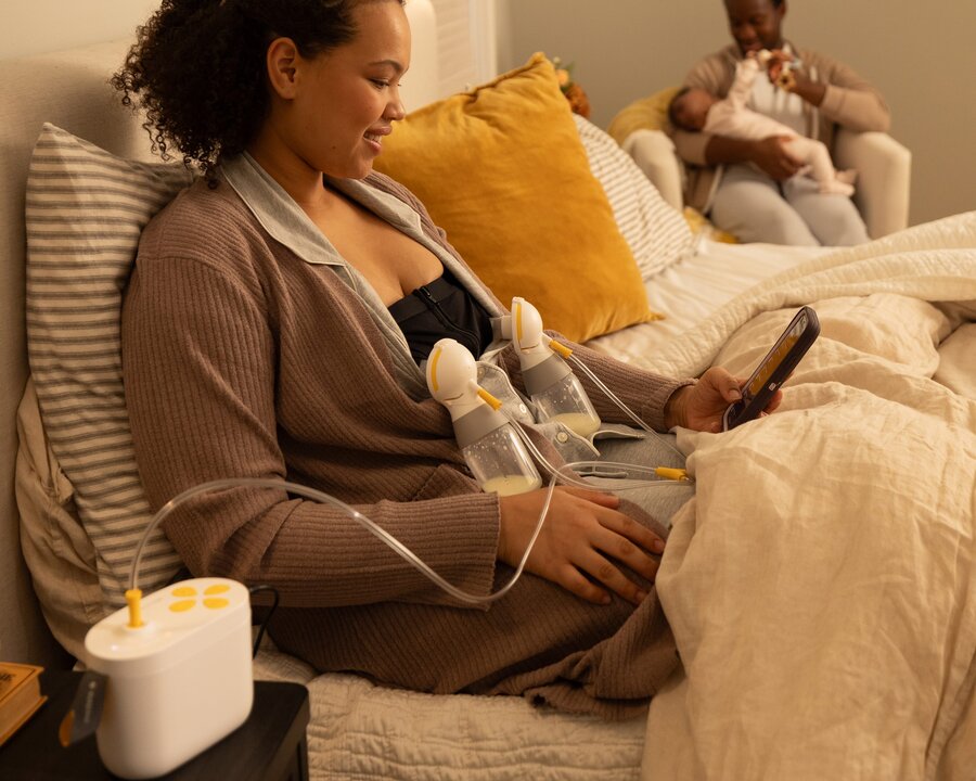 Mom sitting on bed looking at her phone while using Pump In Style Pro breast pump and Hands-free Pumping Bustier.  Dad is playing with baby on a chair in the corner.