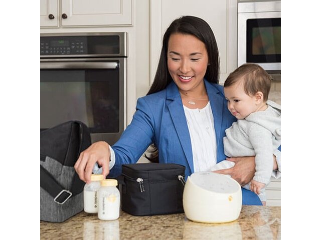 Mom holding baby in kitchen while taking her Medela Sonata® Smart double electric breast pump and storage bottles filled with milk out of her pump bag and cooler