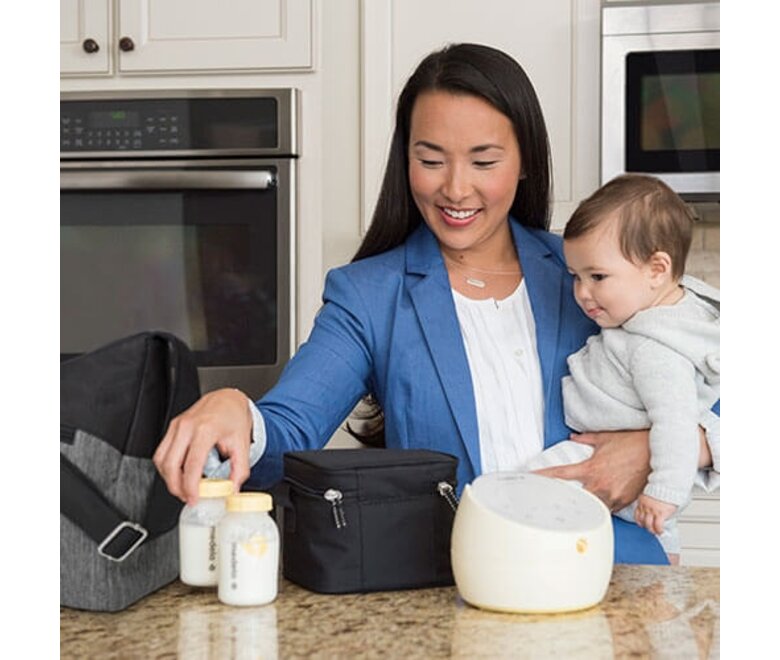 Mom holding baby in kitchen while taking her Medela Sonata® Smart double electric breast pump and storage bottles filled with milk out of her pump bag and cooler