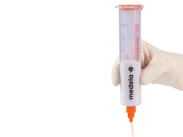 A Medela GraviFeed® Syringe with ENFit® Connector and tubing held by someone's hand wearing a surgical glove.