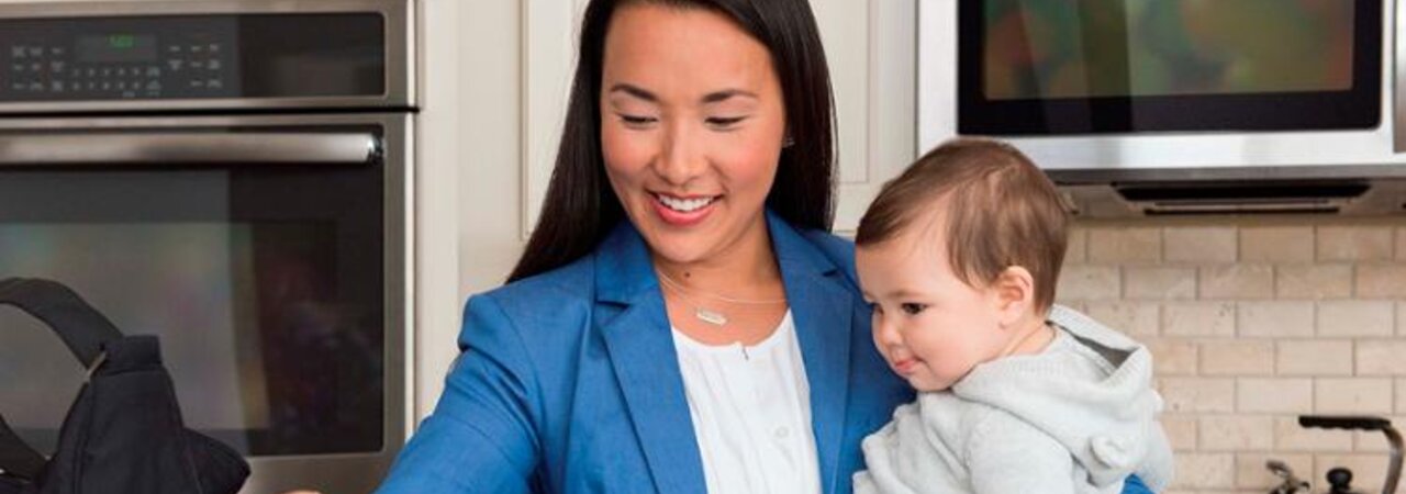 A mom getting ready to go to work while holding her baby on her hip in the kitchen.