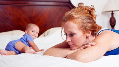 A mom leans against her bed and sighs as her baby crawls and looks back at her.