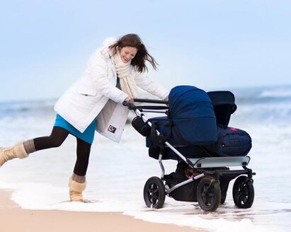 A mom playing in the water on the beach in winter while pushing a double baby stroller.