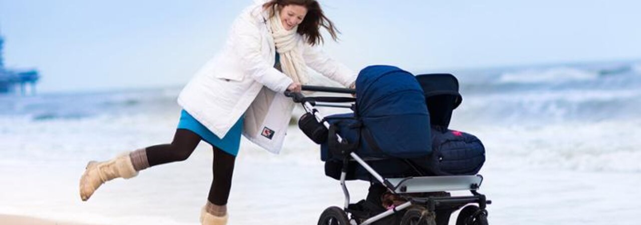 A mom playing in the water on the beach in winter while pushing a double baby stroller.