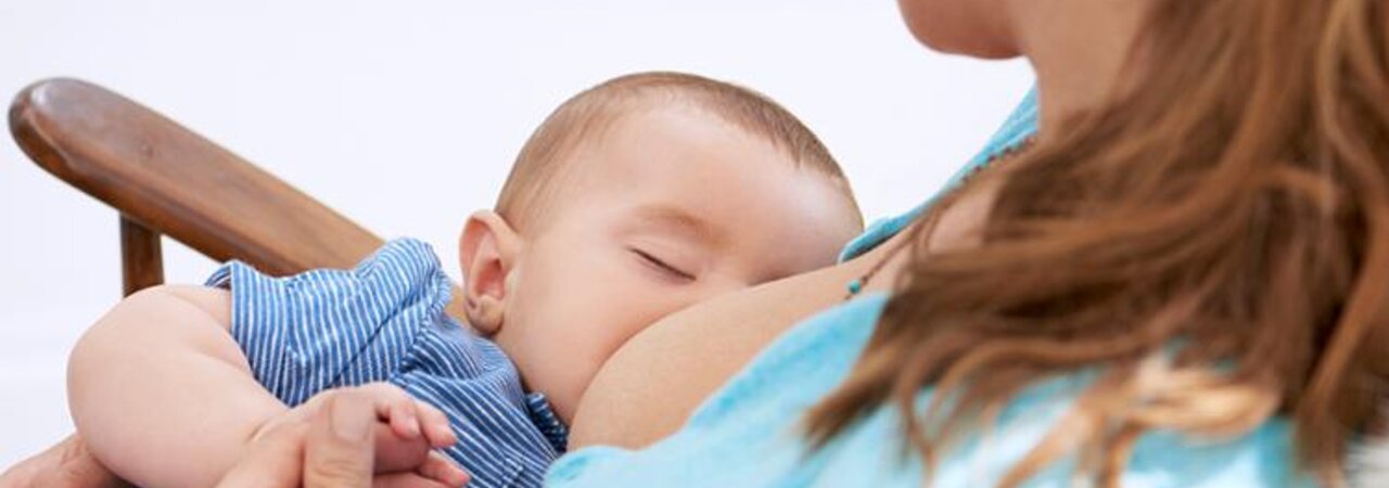 A baby falls asleep while being breastfed in his mother's arms as she sits in a rocking chair.