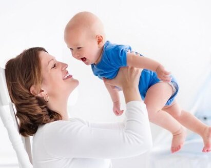 A mother in a rocking chair plays with her baby and laughingly lifts him up and down.