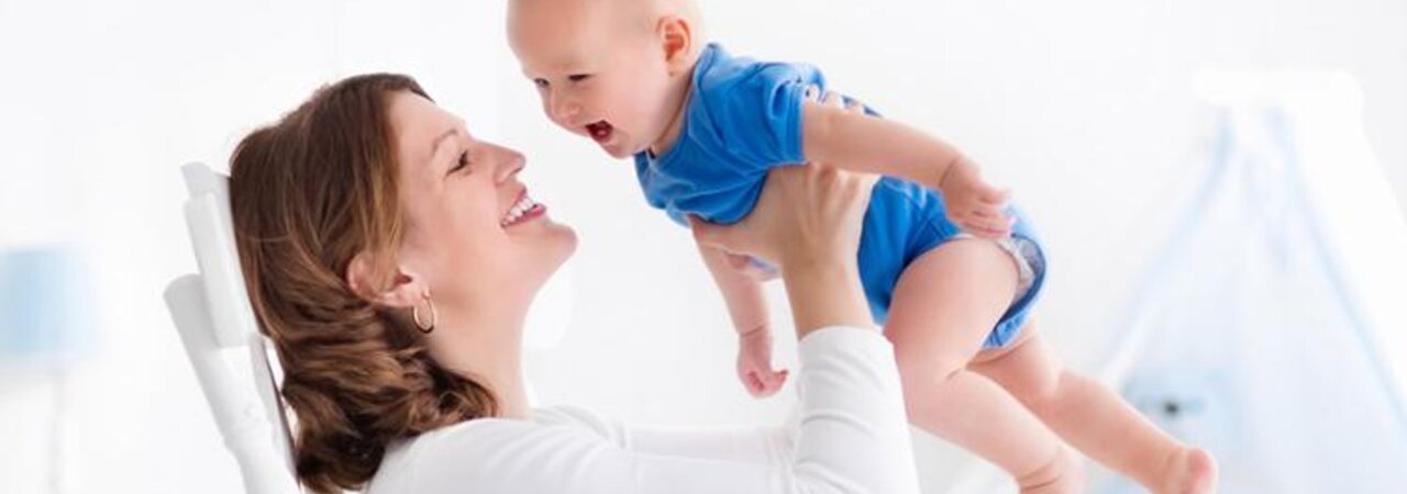 A mother in a rocking chair plays with her baby and laughingly lifts him up and down.