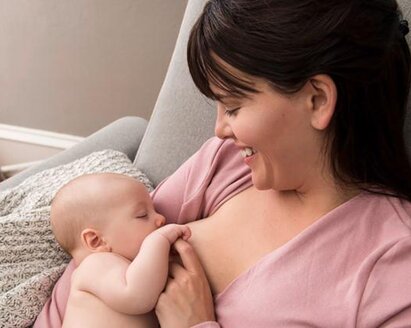 A mother smiles and looks down at her baby as they nurse, sitting at home in the living room.