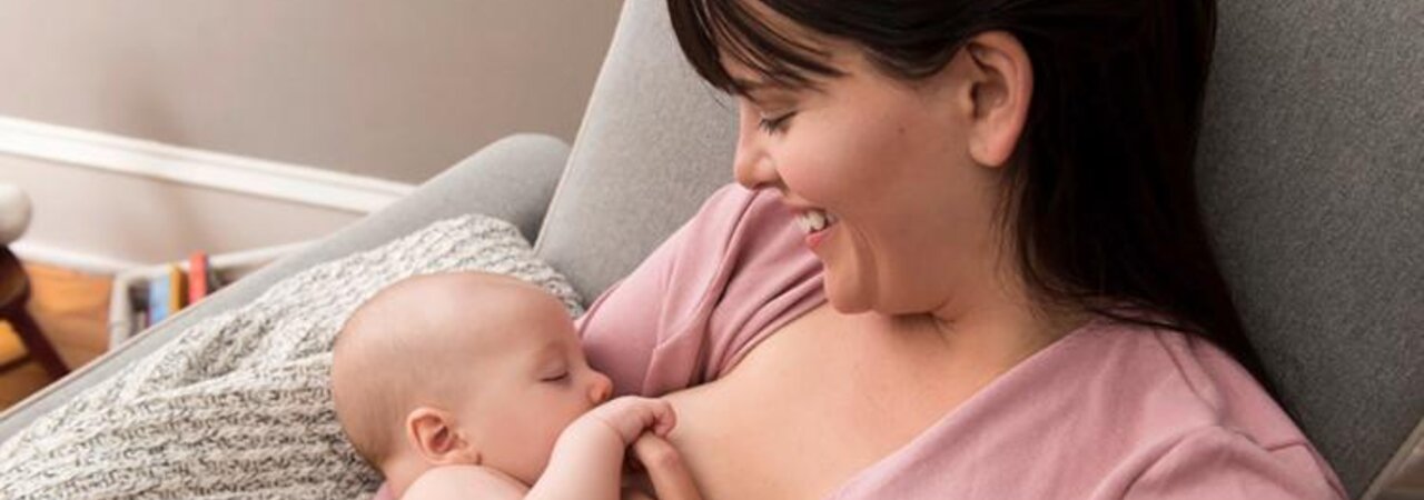 A mother smiles and looks down at her baby as they nurse, sitting at home in the living room.