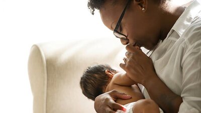 A baby is nursing while its mother kisses its hand as they sit in a chair at home.