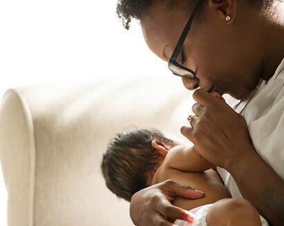 A baby is nursing while its mother kisses its hand as they sit in a chair at home.