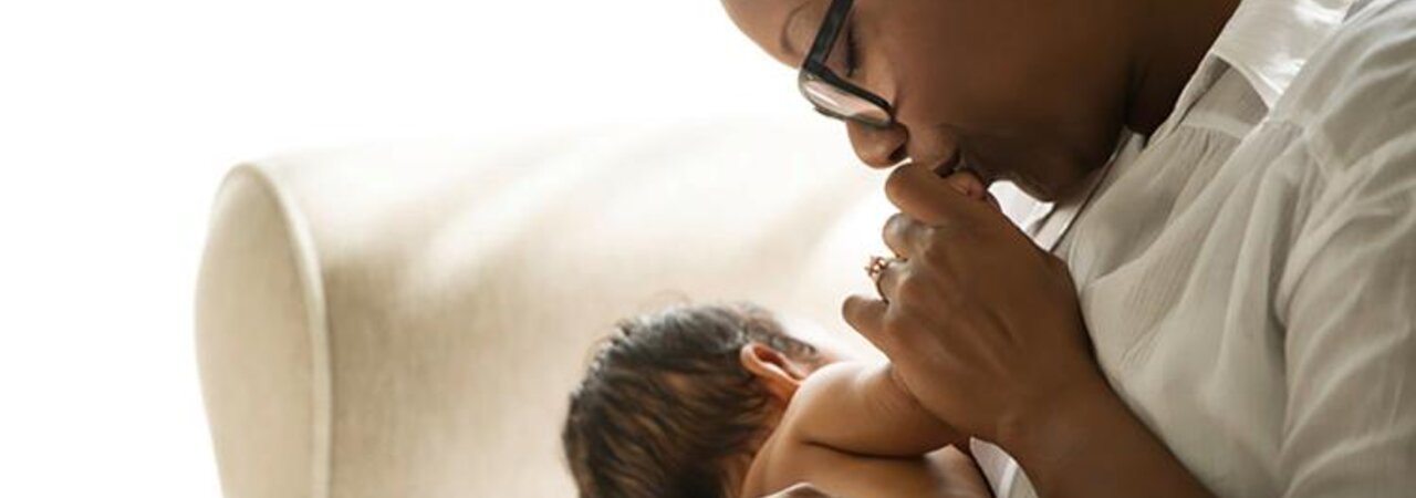 A baby is nursing while its mother kisses its hand as they sit in a chair at home.