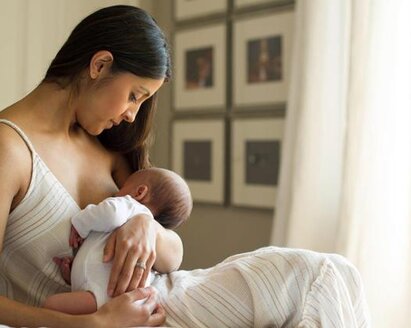 A mother holds her newborn baby while it is breastfed, sitting on the bed near the window at home.