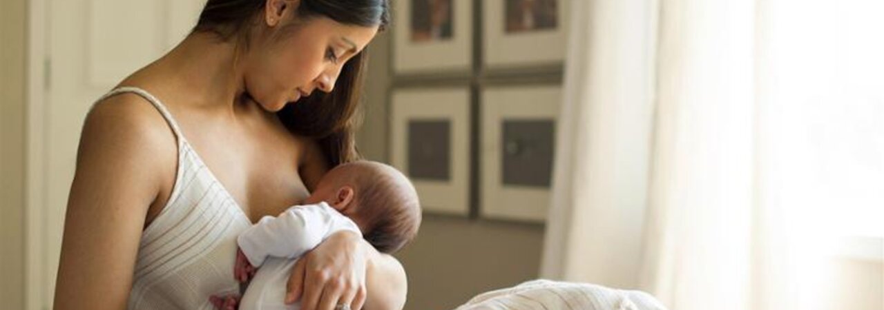 A mother holds her newborn baby while it is breastfed, sitting on the bed near the window at home.