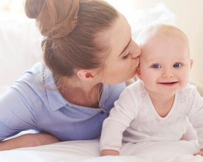 A baby crawls around on the bed while its mother lies next to it and gives it a kiss on the side of the head.