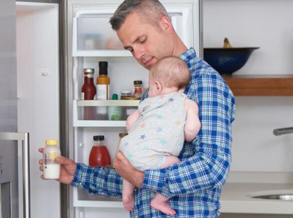 Een vader met zijn baby in zijn armen neemt een fles moedermelk uit de koelkast om op te warmen voor een voeding.