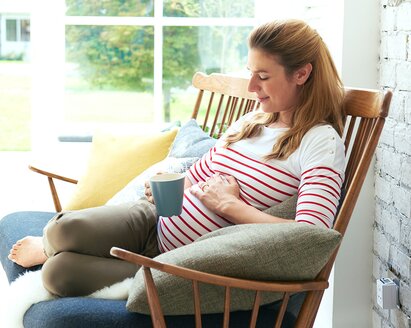 A pregnant woman sits outside on the veranda with a drink in one hand and the other on her pregnant belly.