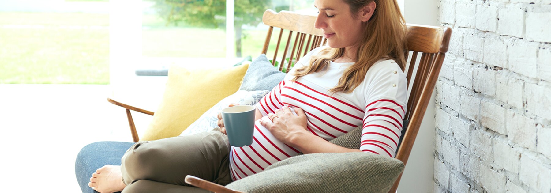 Een zwangere vrouw zit buiten op de veranda met een drankje in haar ene hand en de andere op haar zwangere buik.