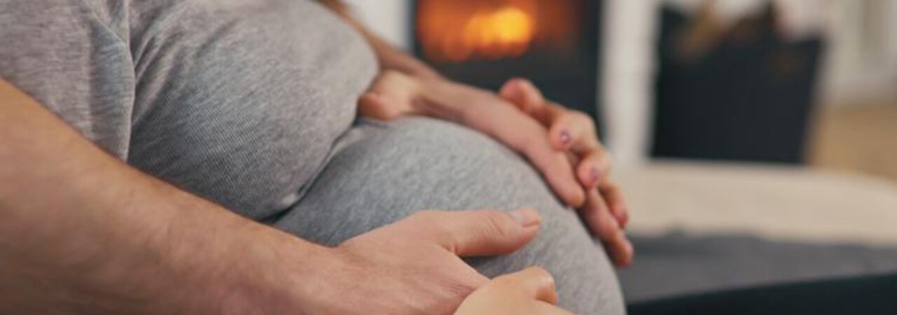 Woman sitting with her hands and her husbands hands on her pregnant belly.