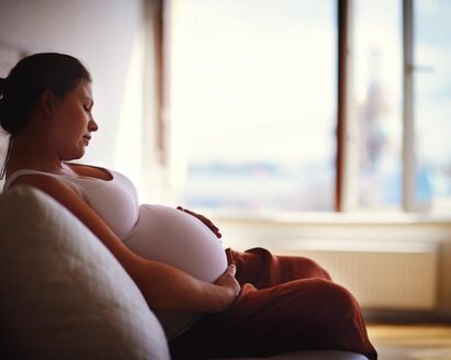 Een zwangere vrouw zit op een bank in een schemerige kamer met haar handen op haar babybump en kijkt tevreden naar beneden.