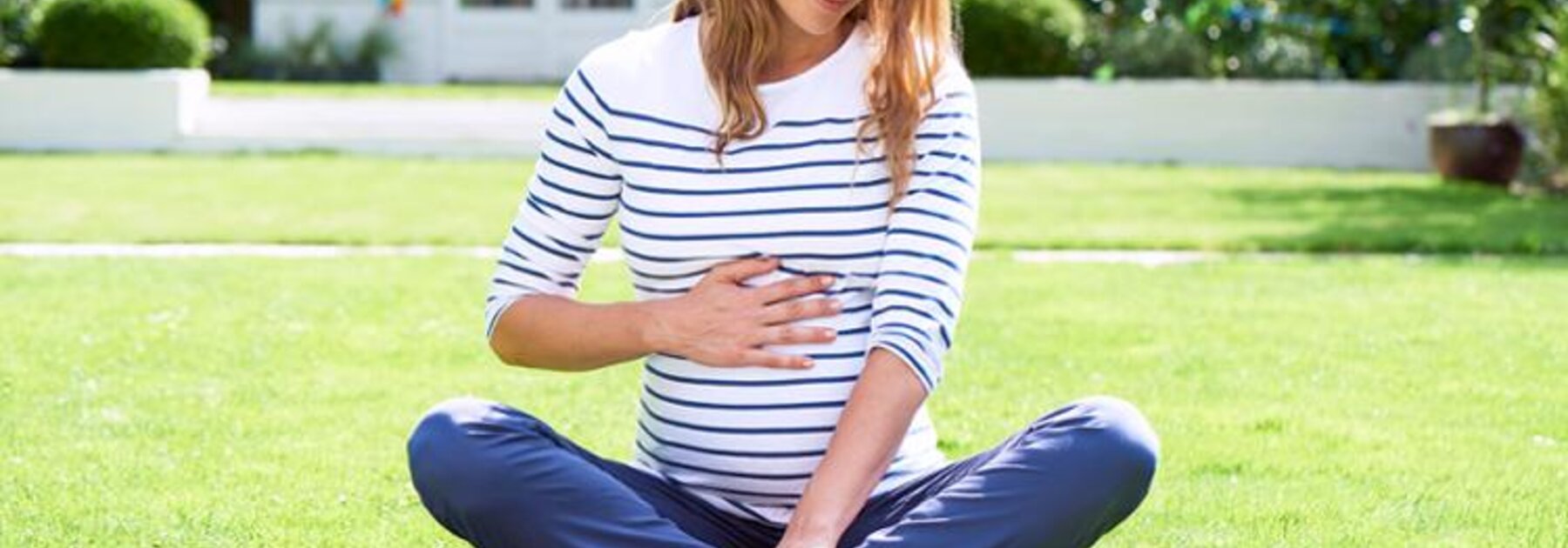 A pregnant woman sitting outside on the grass looking at a book.