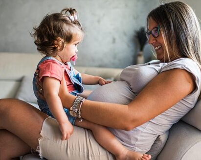 A pregnant woman sits on a couch with her toddler daughter on her lap, facing each other.