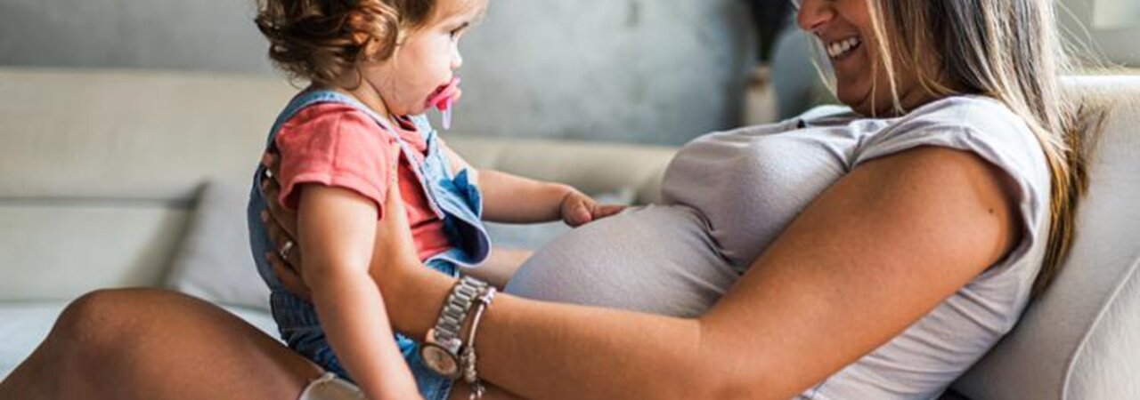 A pregnant woman sits on a couch with her toddler daughter on her lap, facing each other.