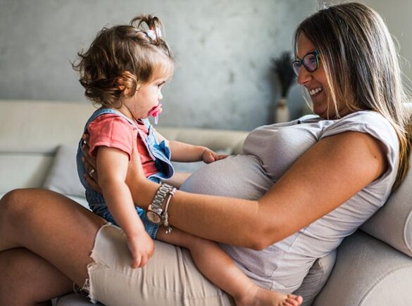 Een zwangere vrouw zit op een bank met haar peuterdochter op schoot, tegenover elkaar.
