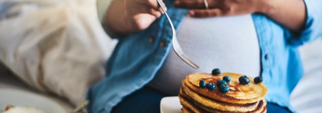 Een zwangere vrouw die haar buik vasthoudt terwijl ze pannenkoeken en fruit eet, zit op een bank.