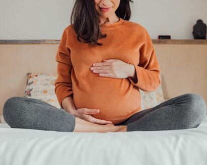 A pregnant mother sits at the end of a bed with her legs drawn up and her hands on her stomach.