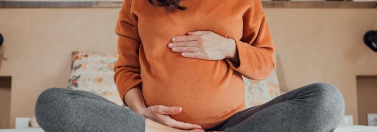 A pregnant mother sits at the end of a bed with her legs drawn up and her hands on her stomach.