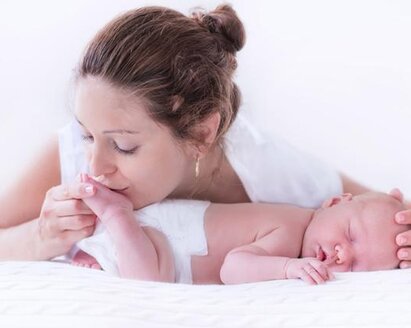 A baby sleeps on its tummy while its mother gives it a gentle kiss on the bottom of its foot and places her hand on its head.