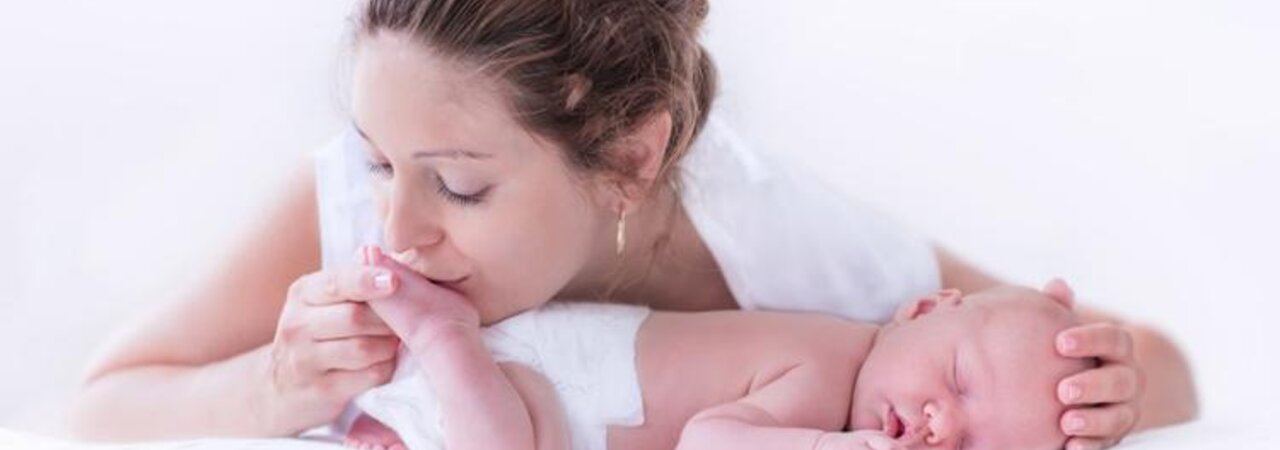 A baby sleeps on its tummy while its mother gives it a gentle kiss on the bottom of its foot and places her hand on its head.