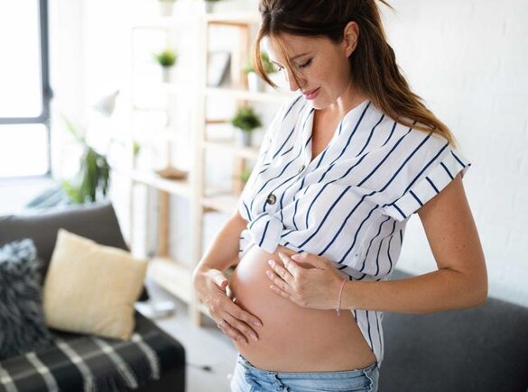 Eine schwangere Frau steht in ihrem Wohnzimmer, legt ihre Hand auf ihren Bauch und spürt, wie sich ihr Baby bewegt.