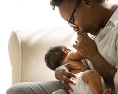 Woman sitting on a couch holding a newborn baby and kissing the baby's hand.