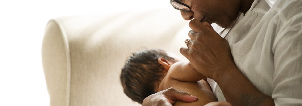 Woman sitting on a couch holding a newborn baby and kissing the baby's hand.