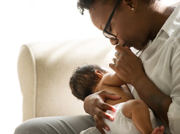Vrouw zit op een bank en houdt een pasgeboren baby vast en kust het handje van de baby.