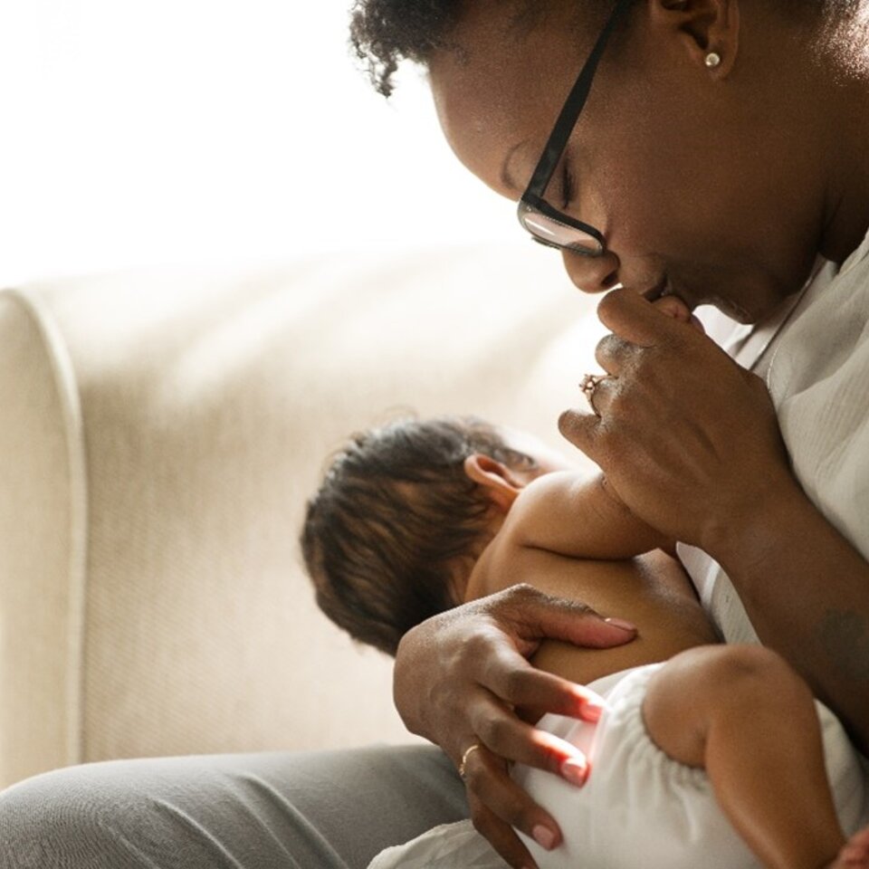 Frau sitzt auf einer Couch, hält ein neugeborenes Baby und küsst die Hand des Babys.