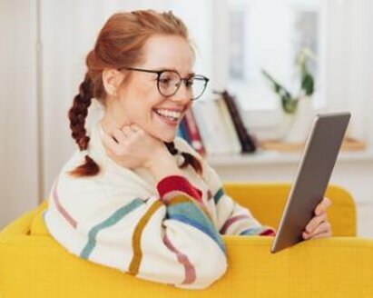 Een vrouw zit thuis op een stoel te lachen en naar haar tablet te kijken.