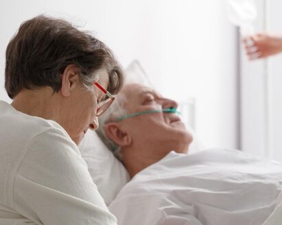 In diesem Krankenhaus sitzt eine Ehefrau bei ihrem sich erholenden Mann, während ein Arzt im Hintergrund seinen Infusionsbeutel anpasst.