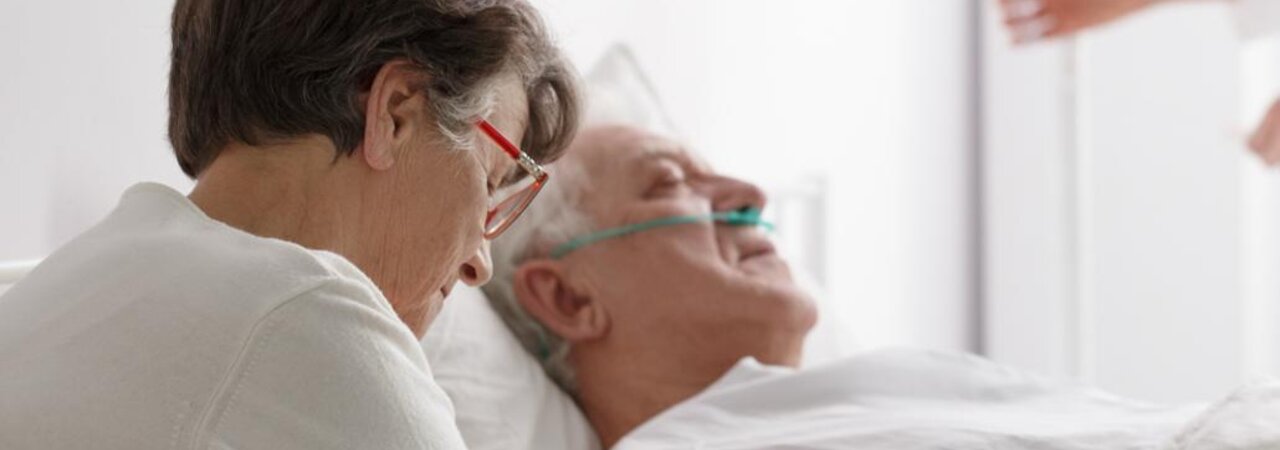 In diesem Krankenhaus sitzt eine Ehefrau bei ihrem sich erholenden Mann, während ein Arzt im Hintergrund seinen Infusionsbeutel anpasst.