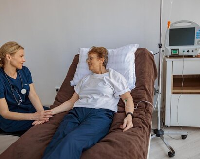 Eine Frau liegt in einem Krankenhausbett, neben ihr sitzt ein Arzt und hält ihre Hand.