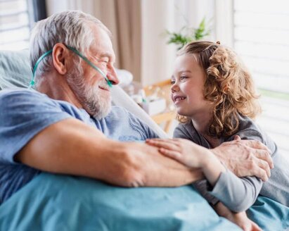 Älterer Mann, der mit einer Nasenkanüle in einem Krankenhausbett liegt und ein junges Mädchen anlächelt, das neben ihm sitzt.