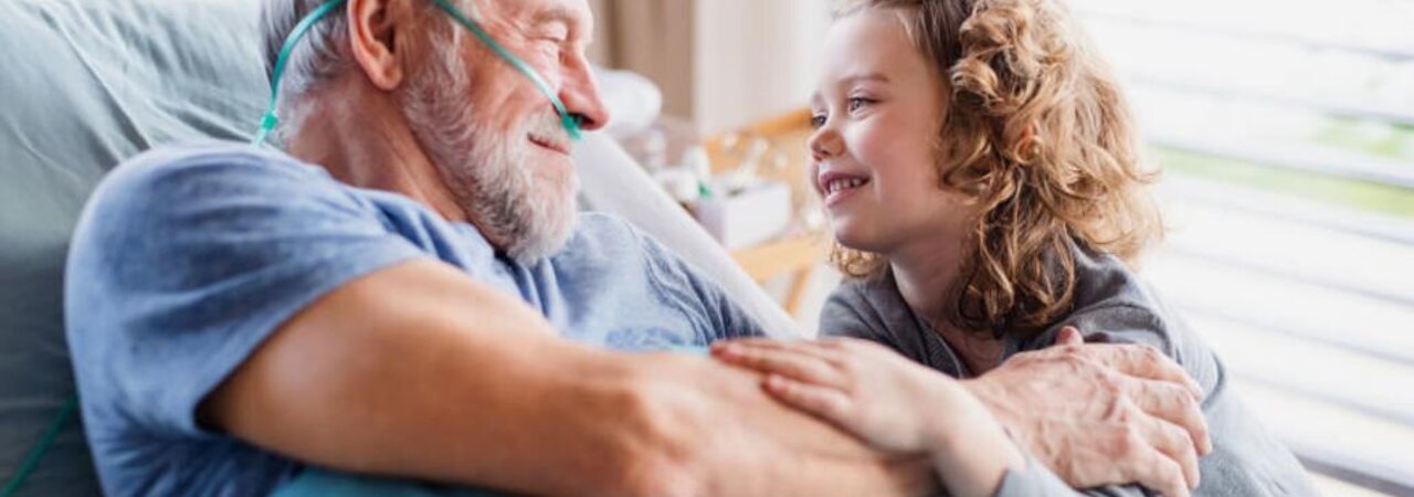 Älterer Mann, der mit einer Nasenkanüle in einem Krankenhausbett liegt und ein junges Mädchen anlächelt, das neben ihm sitzt.