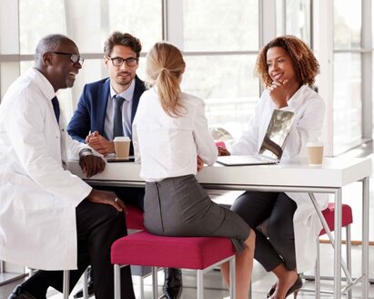 A group of professionals is sitting around a laptop at a table having a discussion.